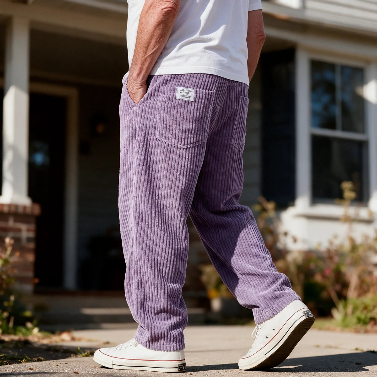 A man walking in front of a house in the Clark pants in purple, with a white T-shirt and white sneakers, one hand in his pocket.